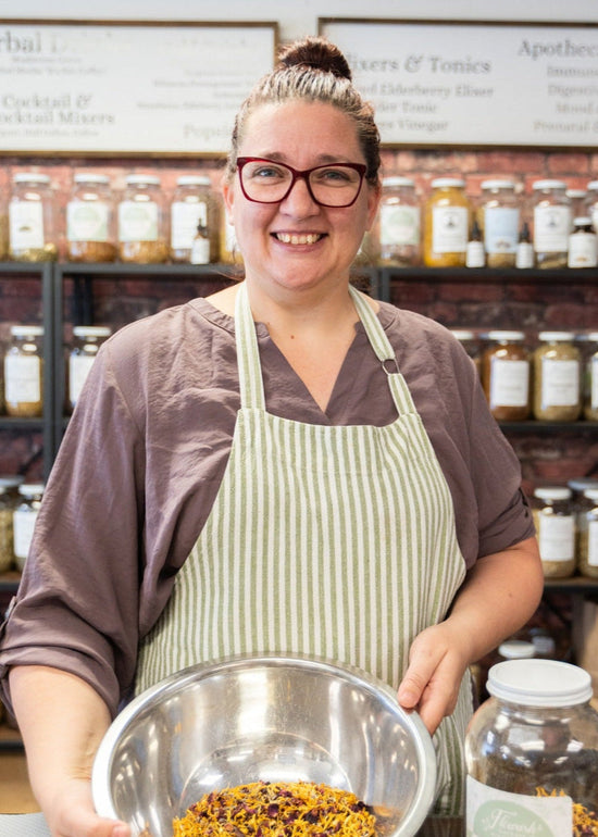 Person holding a bowl of dried herbs in a store with shelves of jars.